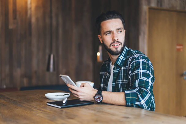 Pensive hipster guy with modern cellphone device thoughtful looking away while creating publication text - Foto, immagini