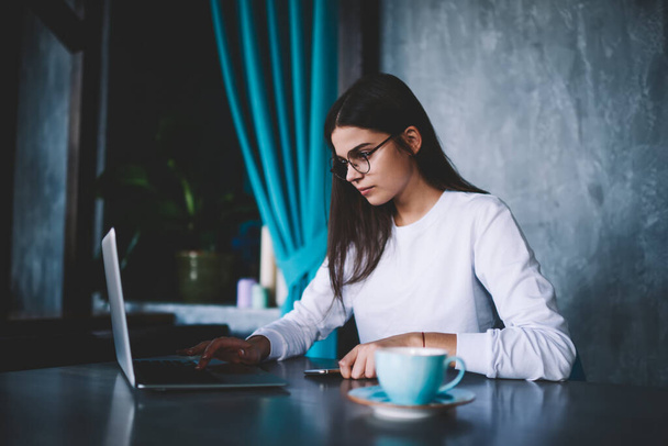 Wistful woman remote worker with eyewear sitting at table with cup of coffee and searching information on laptop during work - Photo, Image