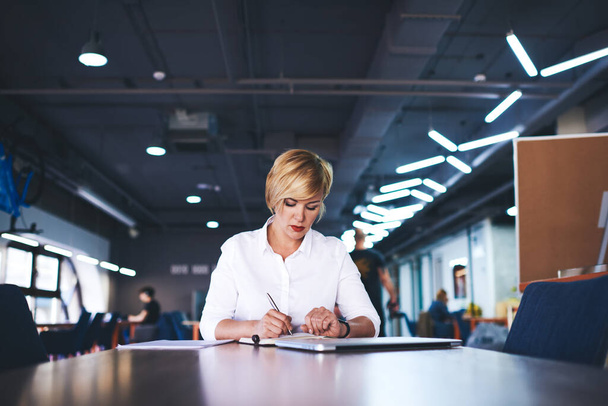 Middle aged female manager looking down and signing documents while sitting at table in modern office and working on project - 写真・画像