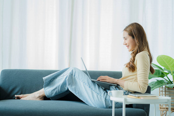 Независимая азиатка, работающая на ноутбуке, проверяет социальные сети, сидя дома на диване и расслабляясь в гостиной. концепция образа жизни для женщин - Фото, изображение