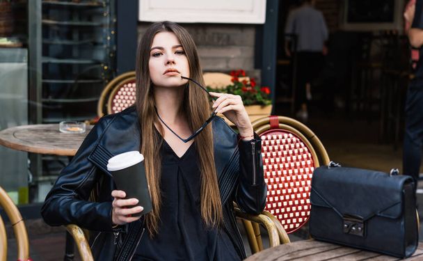 Sensual female model in stylish clothes with long brown hair and trendy handbag holding cup of beverage in one hand and sunglasses - Foto, Bild