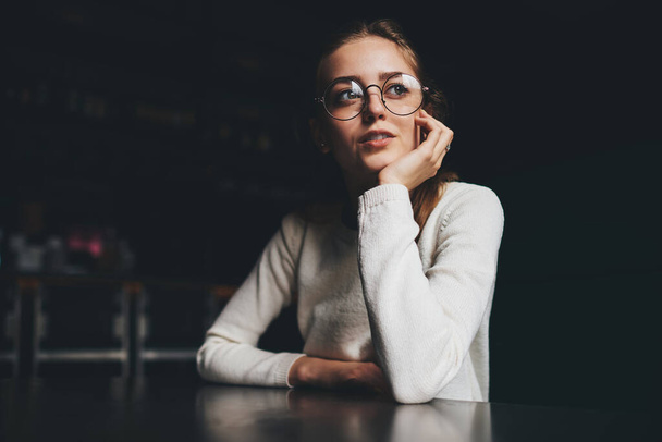 Pensive female in casual white clothes with round eyeglasses looking away sitting at table and leaning on hand in darkness - Foto, Imagem