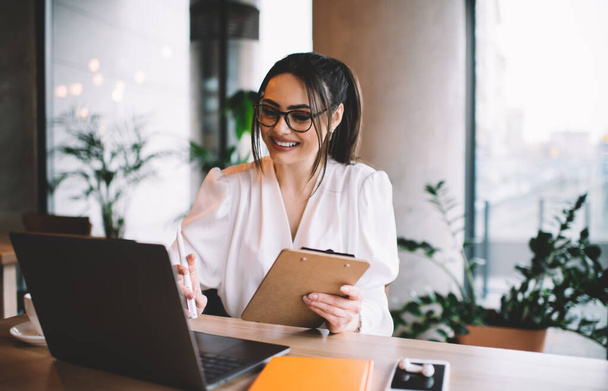 Pozytywna młoda kobieta przedsiębiorca w eleganckiej odzieży z okularami uśmiechnięty i trzymając schowek podczas pracy zdalnie z jasnej przytulnej kawiarni - Zdjęcie, obraz