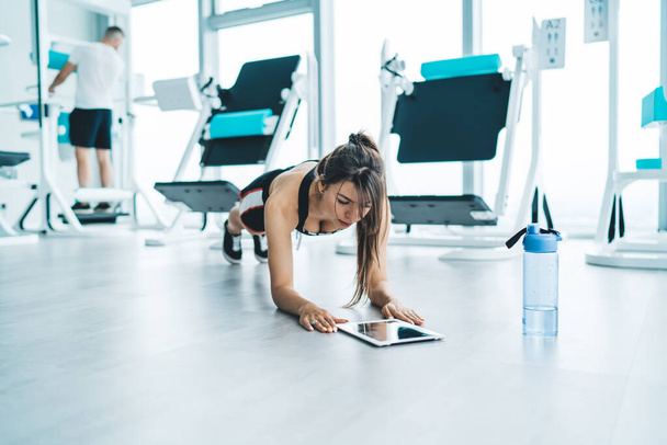 Full length of fit dark haired woman in sportswear and sneakers using tablet while doing plank exercise in modern fitness club - Foto, Imagen
