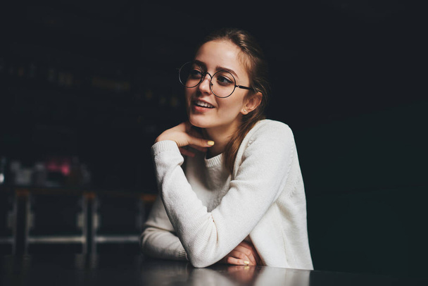 Young female with toothy smile in casual outfit with round frame of eyeglasses looking away sitting at table and touching neck - Фото, зображення