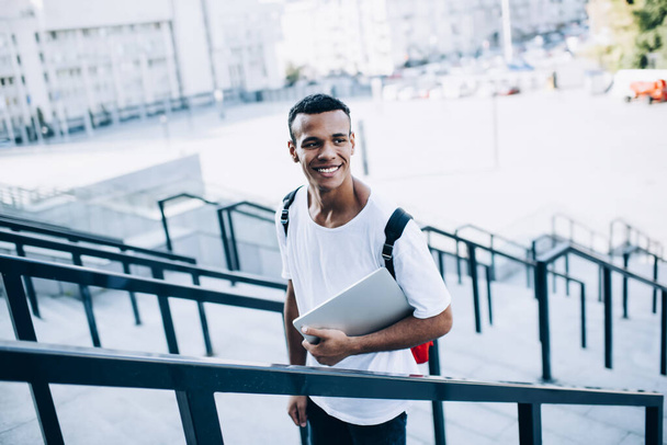 Glad young African American male in casual clothing and with backpack holding modern netbook while standing on concrete stairs with metal railings  - Foto, Imagem