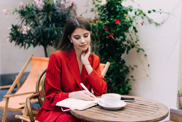 Young female in trendy outfit writing in notebook while sitting at wooden table with cup of hot drink while creating plan in outdoor cafe - Фото, изображение