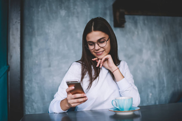 Cheerful woman in casual clothes with long hair and eyewear leaning on hand while sitting at table with cup of coffee and messaging - Foto, Imagem