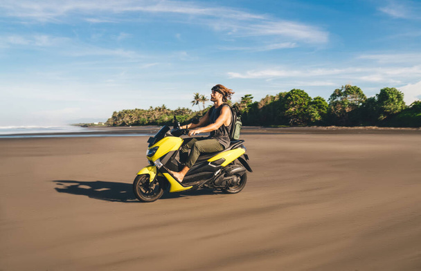 Side view of confident young tanned barefooted male traveler riding fast motorbike on spectacular sandy beach near wavy ocean on sunny day - Foto, Imagen