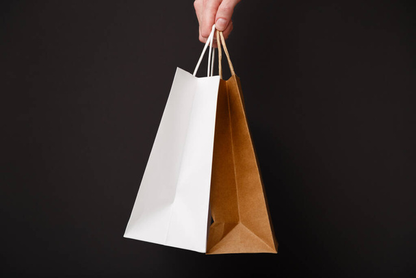 Eine weibliche Hand hält zwei Einkaufstüten isoliert auf schwarzem Hintergrund. Weiße und braune Bastelpapiertüten in der Hand. Black-Friday-Verkauf, Discount, Shopping und Umweltkonzept - Foto, Bild
