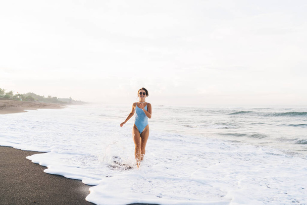 Full body delighted slim female wearing trendy blue swimsuit running on wet seacoast washed by foamy waving sea and looking at camera - Foto, Imagem