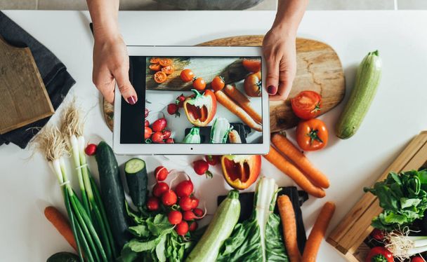 Lähikuva valokuva naisen kädet ottaa kuvan tuoreita vihanneksia digitaalinen tabletti kotona. Pöytälevy näkymä anonyymi nainen tekee terveellisen lounaan tuoreita värikkäitä vihanneksia keittiön pöydällä ja valokuvaus ainesosia hänen tabletti. - Valokuva, kuva