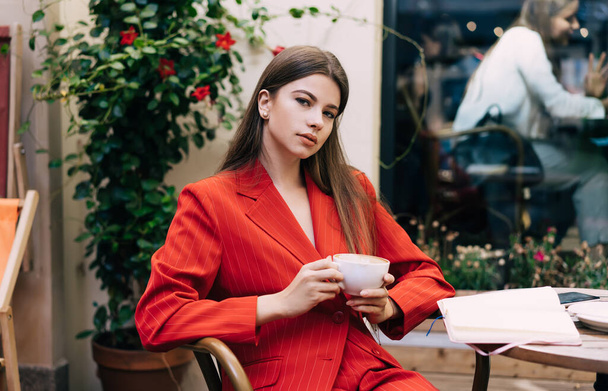 Serious young female in stylish suit sitting at round wooden table with cup of hot drink in street cafe in daytime - Foto, Imagem