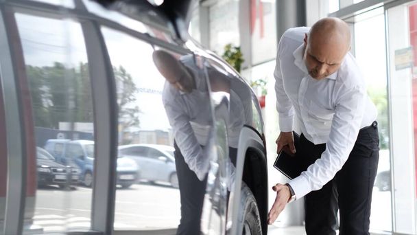 The customer inspects the new car before buying. A bald customer chooses a car at a dealership. People and business concept. - Photo, Image