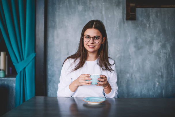 Young cheerful female in casual clothes with eyeglasses looking at camera while sitting at table with cup of coffee during break - 写真・画像