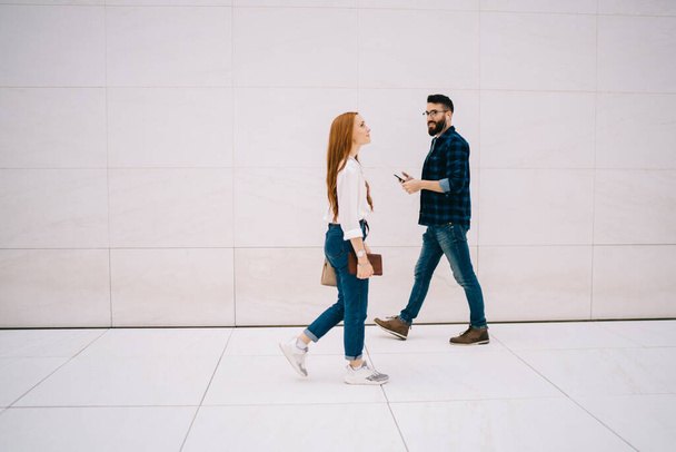 Side view of red hair hipster girl with textbook walking in parallel of Caucasian millennial guy in electronic headphones listening music - Foto, immagini