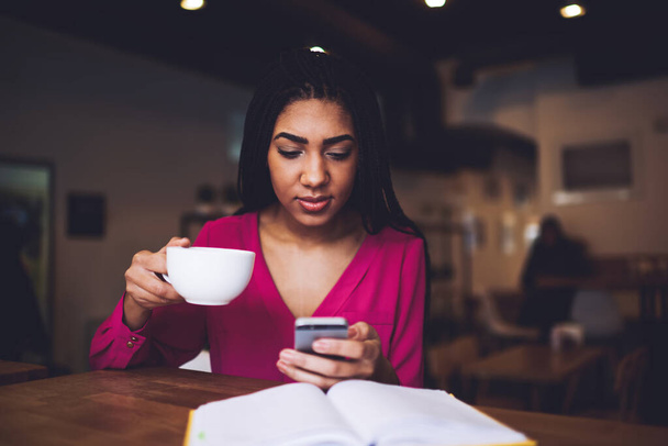 Young wistful African American female sitting at table with cup of hot coffee and surfing mobile phone in light cafeteria - Photo, Image