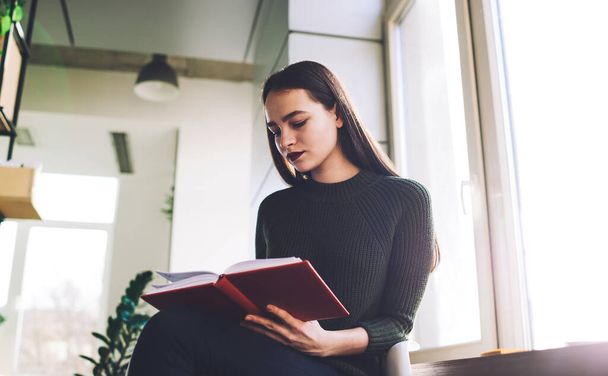 From below of expressive female student in casual outfit sitting near window and reading interesting book with red cover while resting at home - Foto, Imagen