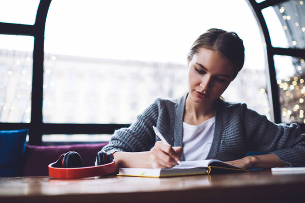Focused woman in casual wear with headphones on table writing in notebook while spending time in cozy cafe near window - Photo, image