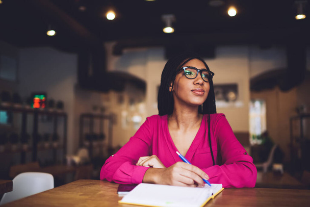 Young thoughtful black female student with eyeglasses looking away while sitting at table with opened notepad doing task in light cafeteria - Foto, Bild