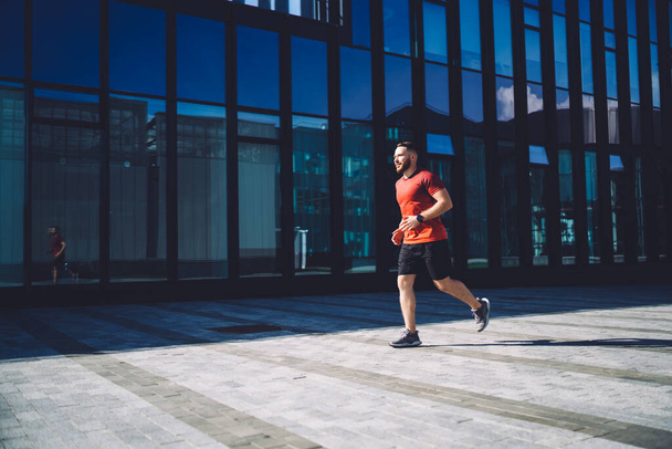 Full length of man in sneakers running on pavement in city downtown against glass building and looking away while enjoying sunny morning - 写真・画像