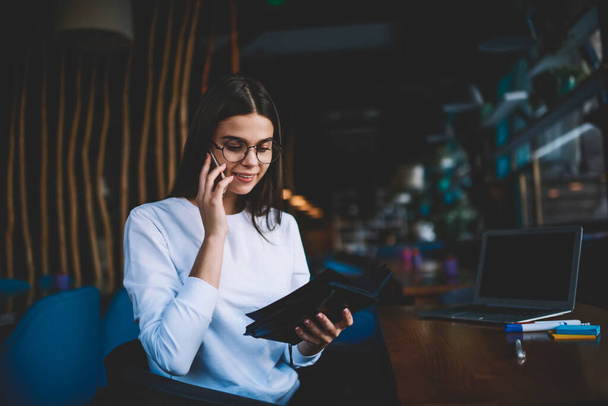 Positive female freelancer in casual clothes sitting at table and reading notes in notepad while having conversation on mobile phone - Foto, immagini
