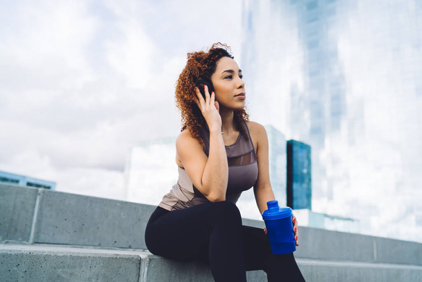 Sporty ethnic woman with bottle of water sitting on stairs in urban environment and listen to music in headphones and looking away - Foto, Imagem