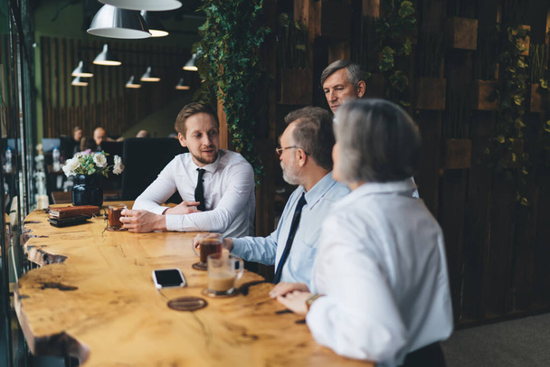 Grupo de colegas en ropa formal sentados en la mesa y tomando bebidas calientes mientras hablan sobre un nuevo plan de negocios en la cafetería - Foto, Imagen