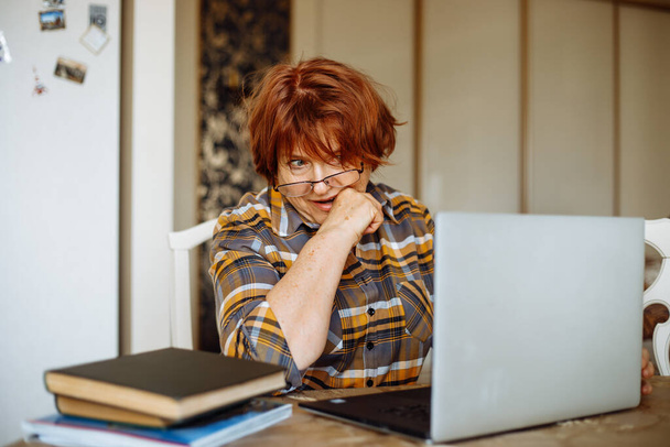 Portrait of senior puzzled nervous woman with dark hair wearing checkered shirt, glasses, sitting at table on white chair, biting finger, looking at laptop, near books, gesturing. Hobby, emotions. - Foto, Bild