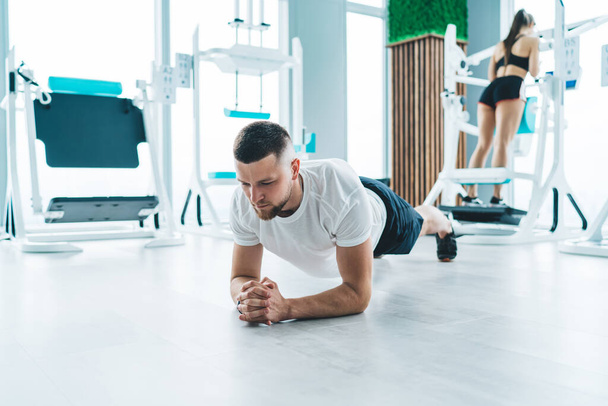 Ernstig gefocust sterke man in activewear doen plank oefening op de vloer in helder daglicht in ruime fitnessruimte met metalen fitnessapparatuur - Foto, afbeelding