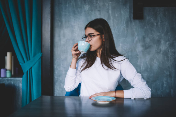 Young thoughtful brunette with long hair and eyewear looking away while sitting at wooden table with saucer and drinking hot coffee - Valokuva, kuva