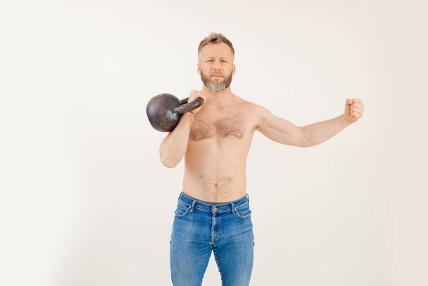 Portrait of middle-aged muscular shirtless bearded man with fair hair wearing blue jeans, lifting kettle bell, showing muscles, standing on white background. Sport, weightlifting, workout, fitness. - Foto, Imagem