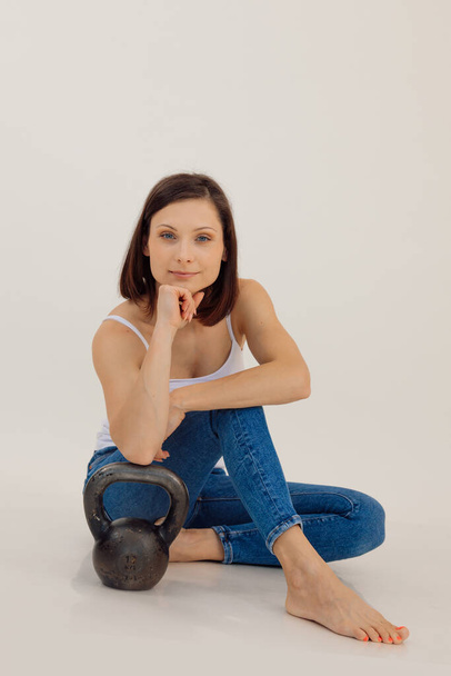 Portrait of young slim sporty muscular woman with dark hair wearing white top, blue jeans, sitting, leaning on kettle bell, putting chin on hand. Sport, strength, workout, fitness. Studio, vertical. - 写真・画像