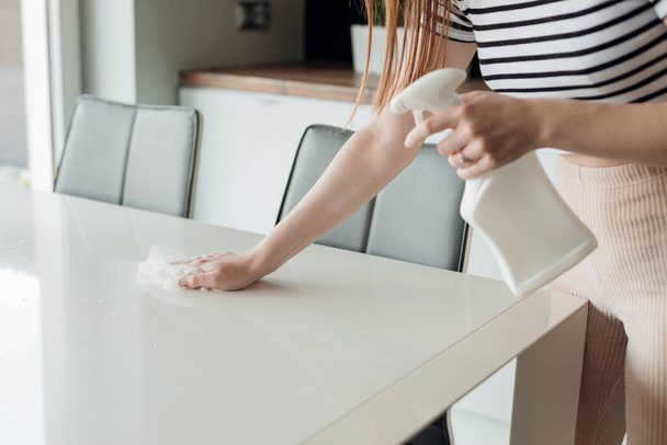 Eine unkenntliche Frau steht in der Küche, sprüht aus einer Pulverflasche auf einen weißen Tisch, wischt mit Lappen, desinfiziert die Reinigung, desinfiziert die Oberfläche. Hausarbeit, Hausarbeit, COVID-19 Prävention. - Foto, Bild