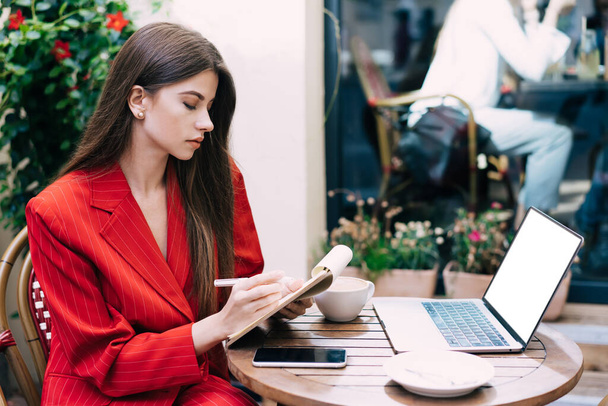 Side view of pensive attractive businesswoman with notebook and pen in hands sitting at table with laptop and cup of coffee against window - Foto, Imagem