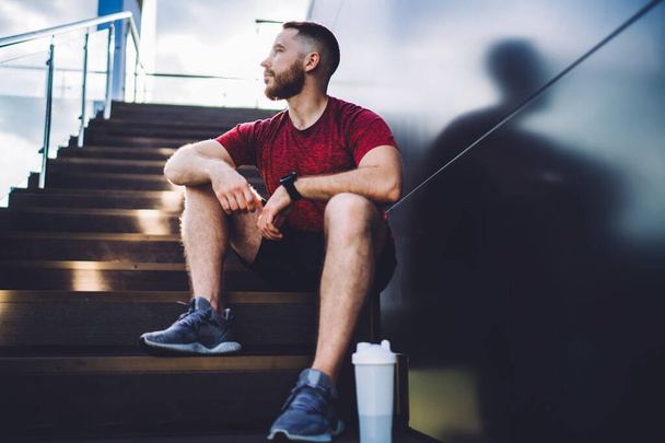 Low angle of bearded male with smartwatch sitting near metal wall and looking away during break from training in street - Foto, afbeelding