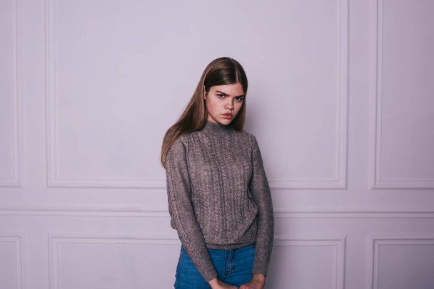 Young serious female in casual sweater and jeans with long hair looking at camera while standing in light room with white wall - Fotó, kép