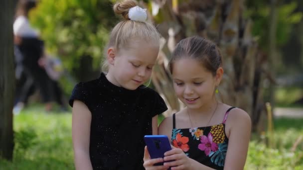 Dos niñas hermanas sentadas en el césped y jugando con el teléfono. Mid shot - Metraje, vídeo