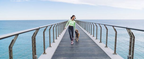 Happy Caucasian woman in sportive tracksuit running at boardwalk way during playful game with morgel dog - 写真・画像