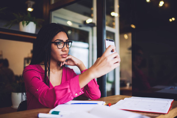 Elegant African American lady with braids in casual wear and spectacles taking selfie on smartphone while working with notes in cafe - Foto, Bild