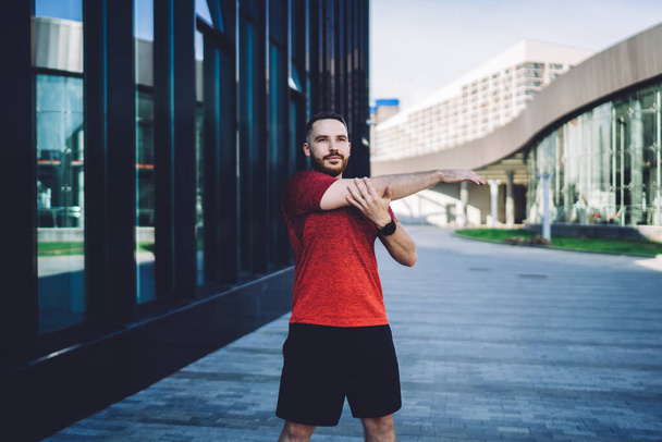 Positive bearded male in casual wear standing alone on pavement and stretching body while exercising among modern buildings during morning and looking away - Photo, Image