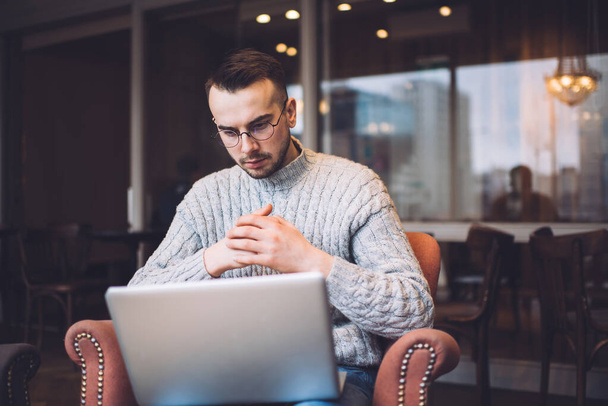Focused bearded male remote worker in casual outfit and eyeglasses sitting in comfortable armchair and surfing laptop during work on project - Photo, Image