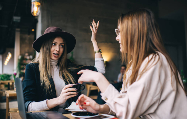 Modern bayan arkadaşlar konuşurken ve el kol hareketleri yaparken heyecan verici haberleri paylaşıyorlar ve modern kafeteryada buluşmalarında kahve içiyorlar. - Fotoğraf, Görsel
