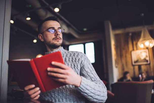 Serious young bearded male in casual clothes and eyeglasses sitting in comfortable armchair in cafeteria and looking away while reading interesting book - Fotó, kép