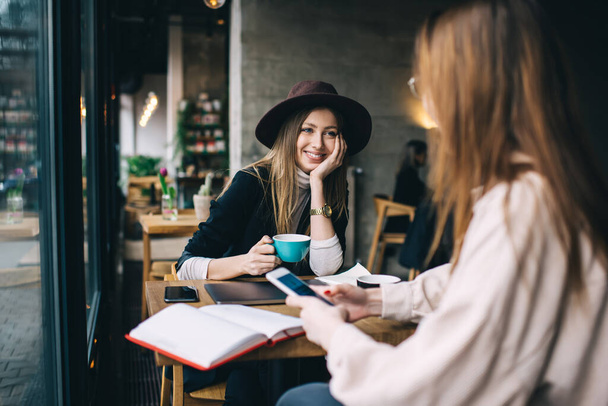 Cheerful female friends in trendy clothes sitting at wooden table and having conversation about plans while enjoying hot coffee together - Fotoğraf, Görsel