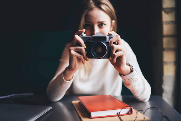 Selective focus of vintage photo camera in hands of young female photographer sitting at table with laptop and book looking at camera - Photo, image
