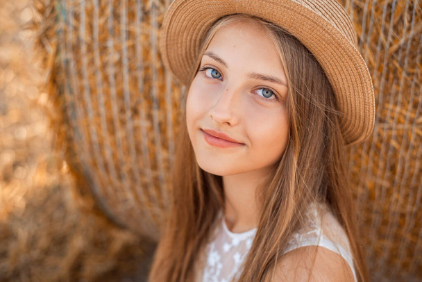 Close-up portrait of a young girl in a light white dress and a hat near the straw bale - Foto, Bild