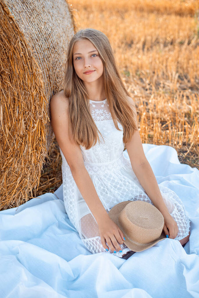 Lovely blond girl sitting under the bale of straw on a white bedsheet holding straw hat in her hands - Foto, imagen