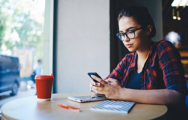 Side view of concentrated on screen ethnic lady in plaid shirt and eyeglasses with smartphone in hands and notebook and tablet on table working - Foto, imagen