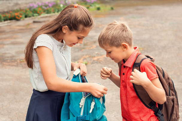 Deux écoliers avec des sacs à dos se tiennent à l'extérieur sur l'aire de jeux près de l'école. Les deux regardent curieusement dans le sac de la fille. Amitié, vie scolaire. - Photo, image